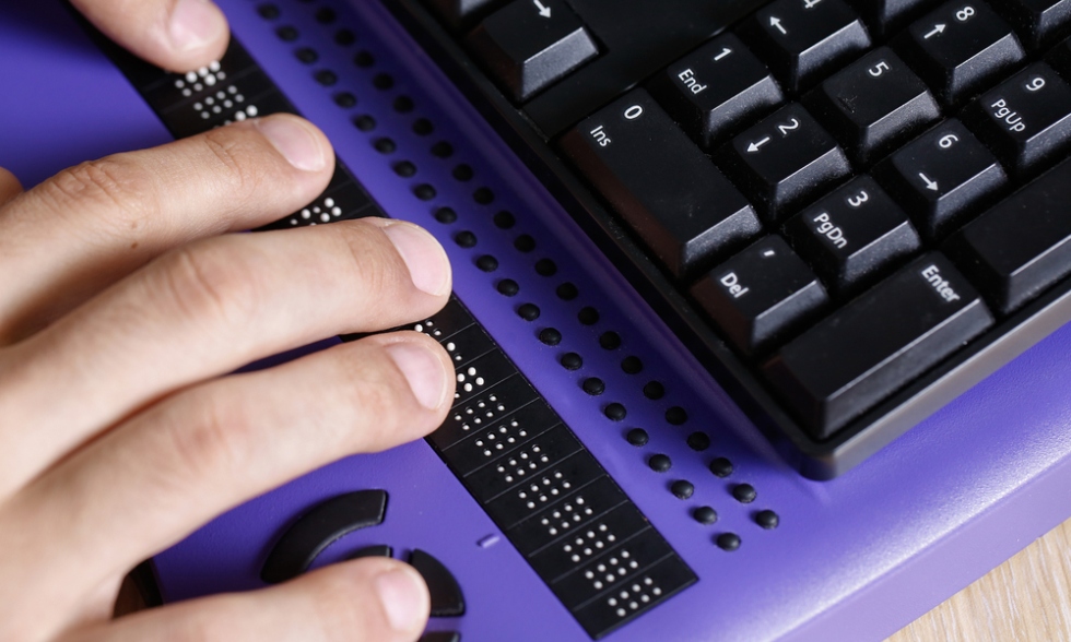A hand is placed on top of a laptop, just below the keyboard where braille is located. The laptop is purple with black keyboard keys.