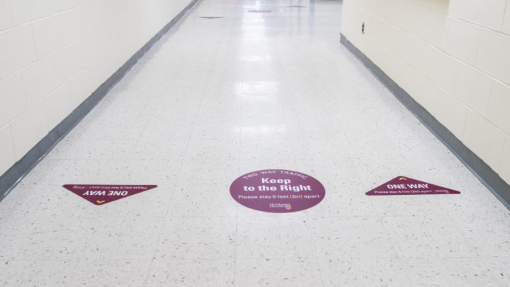 Signs pasted onto a while floor which idenitify how students walking forward need to walk to their right side of the hallway. The sign in the middle explains that this is to stay 6 feet (2) apart due to the COVID-19 pandemic.
